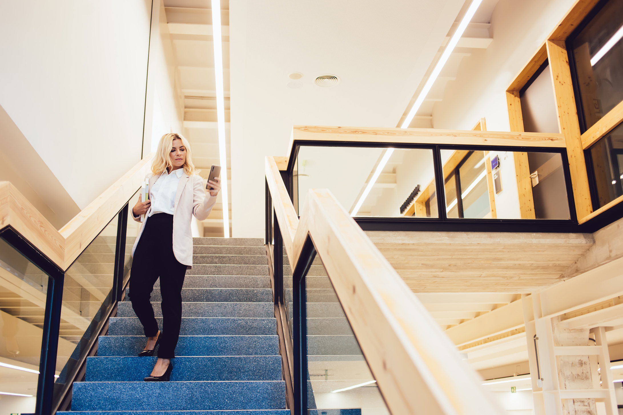 Adult student with literature books going down on university stairs checking information on modern cellular gadget, Caucasian smart casual woman with education textbook browsing school website